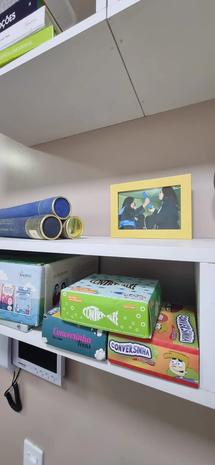 Estante com livros, jogos terapêuticos e foto da Jayane em atendimento